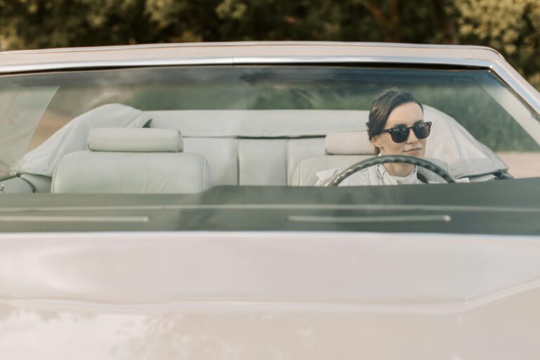 A woman in sunglasses driving a convertible car outdoors on a sunny day.