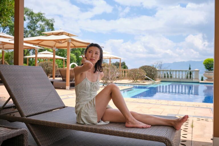An adult woman in a summer dress relaxes on a lounge chair by a resort pool under a sunny sky.