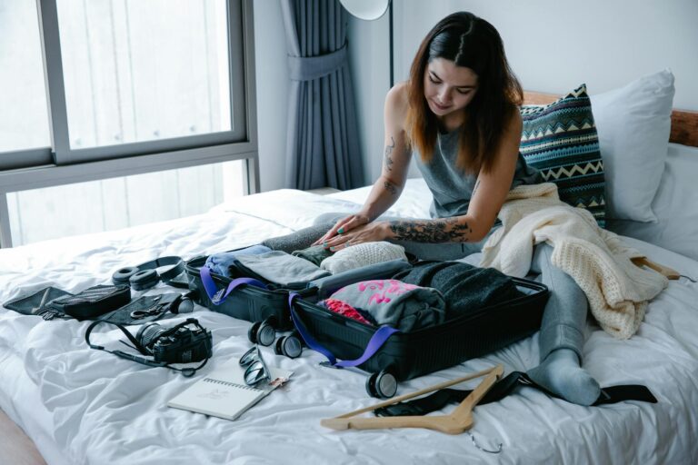 Woman sitting on bed packing suitcase with clothes and travel essentials.